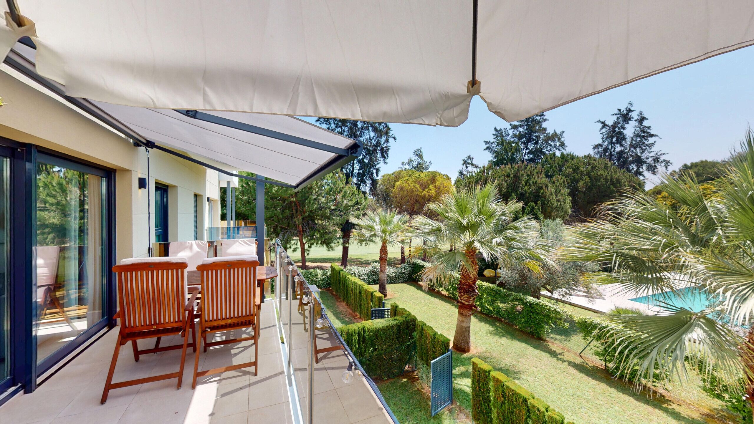 Un balcón con dos sillas de madera y una mesa, con vista a un jardín cuidado que incluye diversas plantas y una piscina.