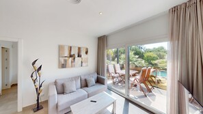 Een moderne woonkamer met een grijze bank, een bijzettafeltje en een schilderij aan de muur. Er is een glazen deur die leidt naar een buitengebied met een zwembad en stoelen.