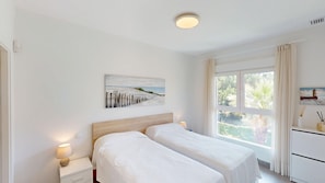 Ein Schlafzimmer mit zwei Betten, einem Bild, einem Fenster mit Vorhängen und einem Nachttisch mit Lampe.