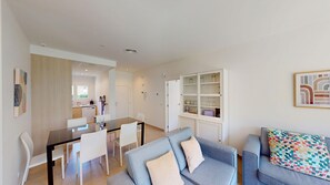 Ein modernes Wohnzimmer mit Essbereich, einem grauen Sofa, einem farbenfrohen Kissen und einer weißen Kommode mit Glastüren.