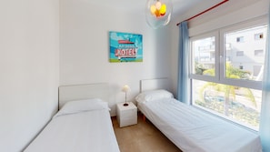 Quarto de hotel com duas camas de solteiro, mesa de cabeceira com luminária, janela com vista para prédios e um aviso de 'VACANCY' na parede.