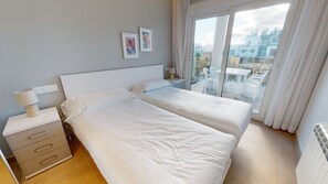 A hotel room with two beds, a bedside table with a lamp, and a view of the city through a large window.