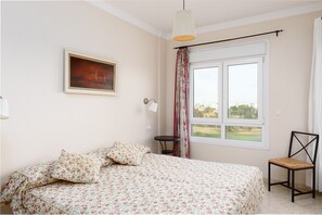 Une chambre à coucher avec un lit, une chaise, une petite table, une lampe, une fenêtre et un tableau accroché au mur.