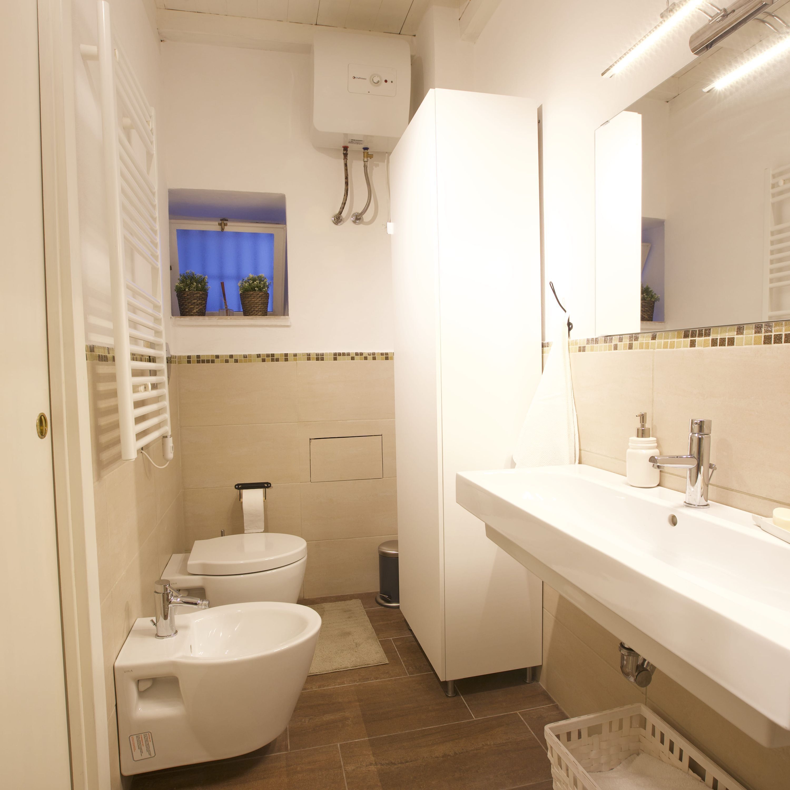 Un baño con inodoro, bidé, lavamanos y una ventana con plantas.