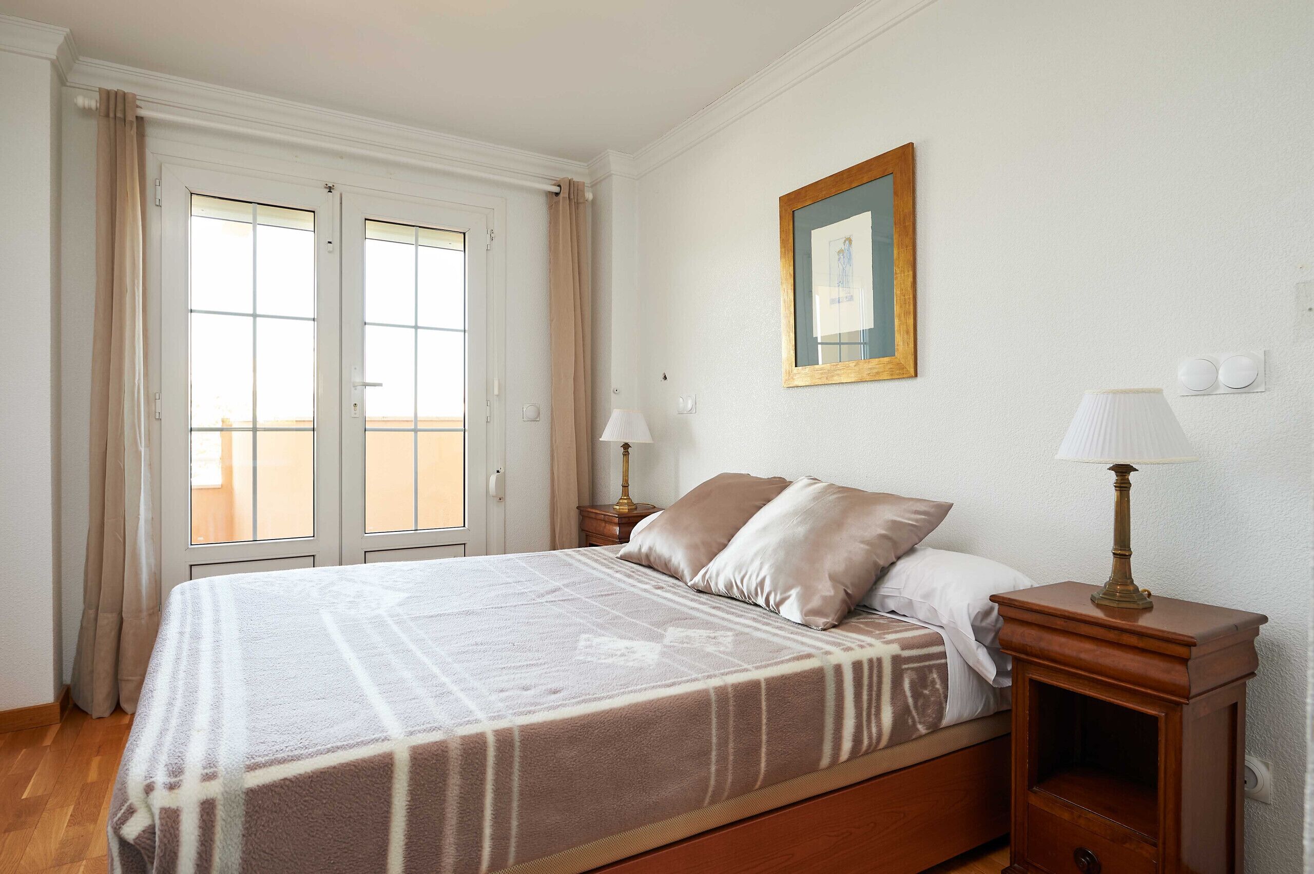 A bedroom with a bed, two bedside tables with lamps, a framed picture on the wall, and a window with curtains.