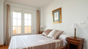 Un dormitorio con una cama, dos mesitas de noche con lámparas, un cuadro enmarcado en la pared y una ventana con cortinas.