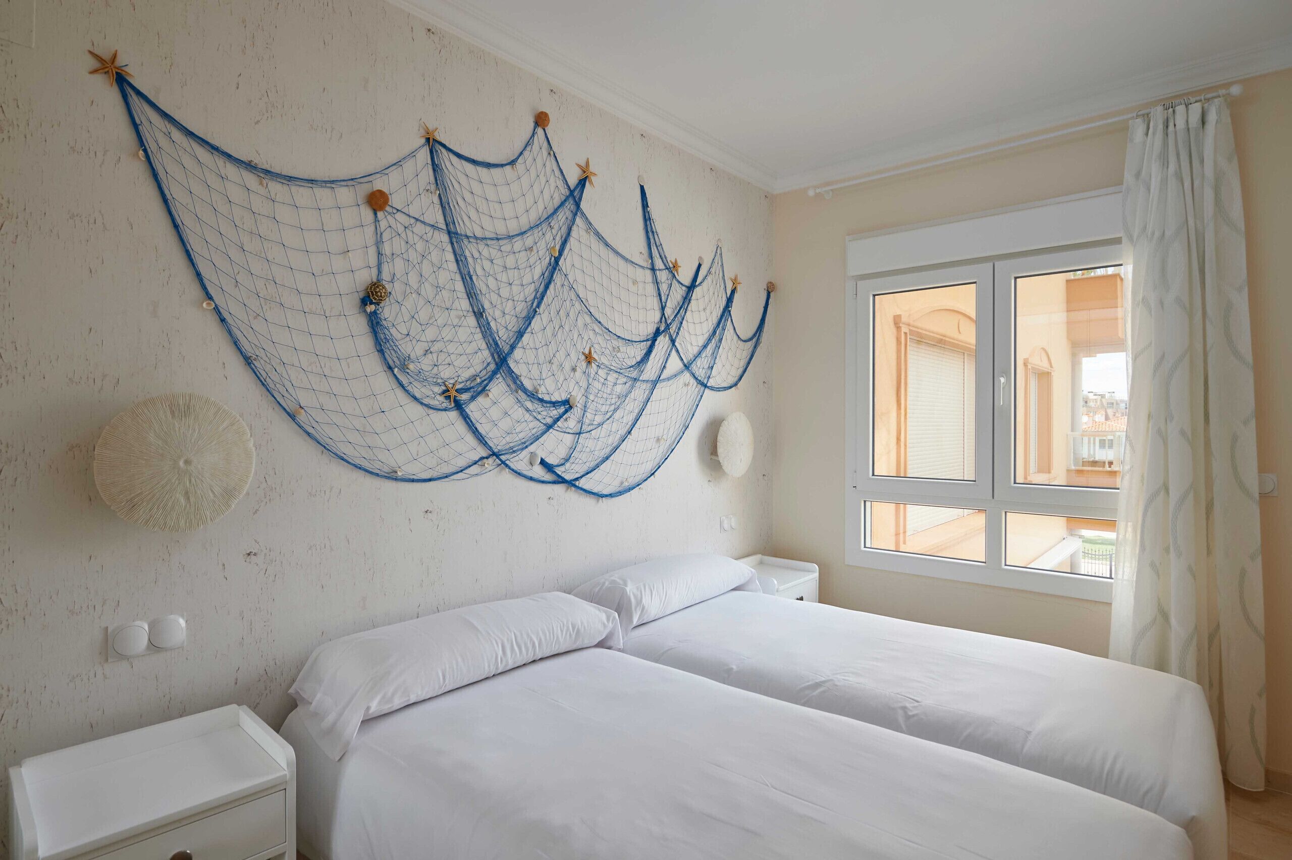 Une chambre avec deux lits, une grande fenêtre avec des rideaux et des éléments décoratifs en forme de filets tendus au mur.