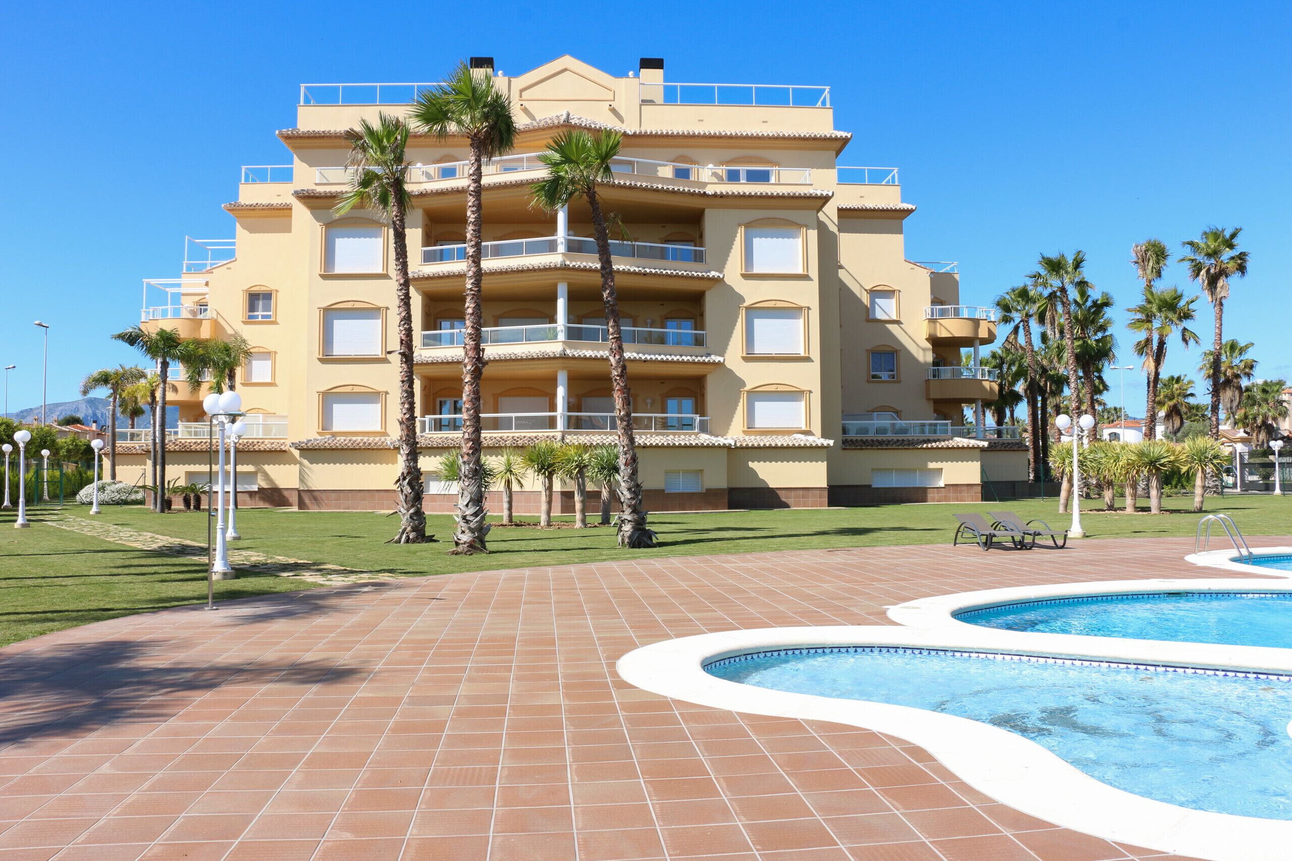 A multi-story hotel with a swimming pool, palm trees, and a clear blue sky.