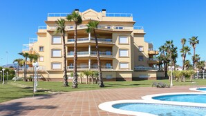 A multi-story hotel with a swimming pool, palm trees, and a clear blue sky.
