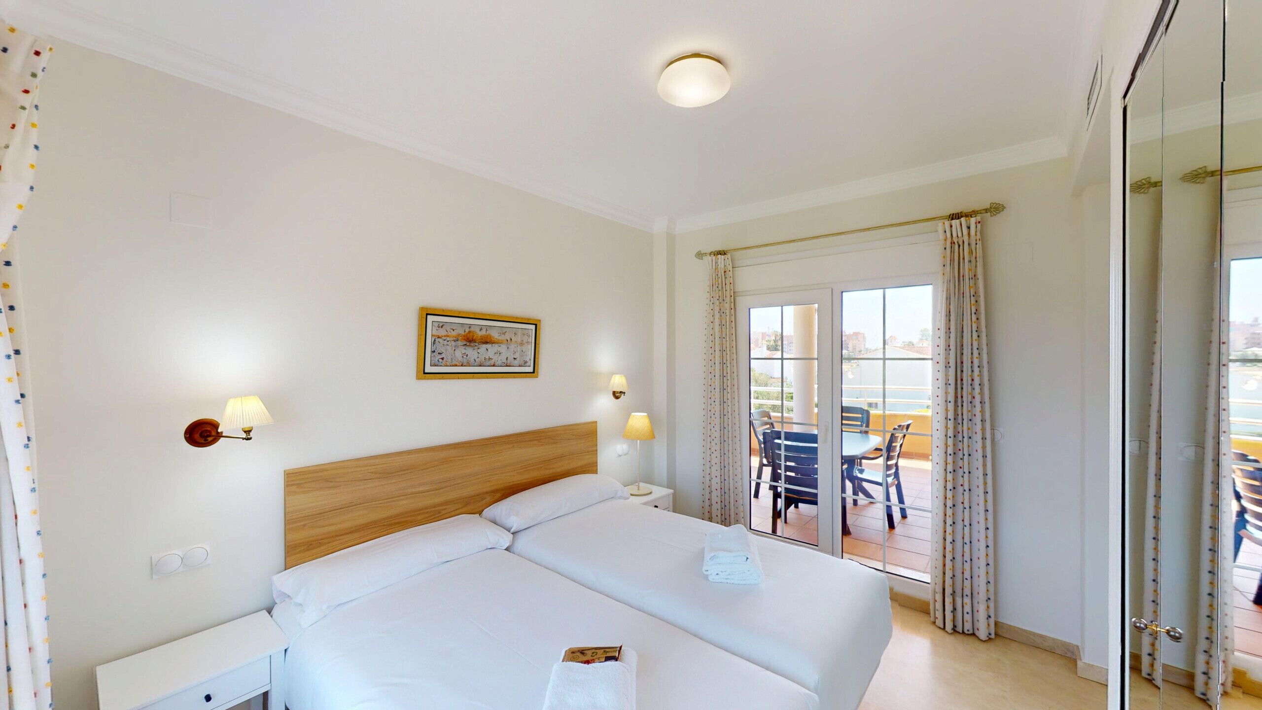 A hotel room with a bed, bedside lamps, a painting, and a balcony with chairs.