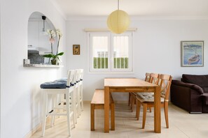 Un coin repas avec une table et des chaises en bois, un banc blanc, une plante en pot et un tableau accroché au mur.
