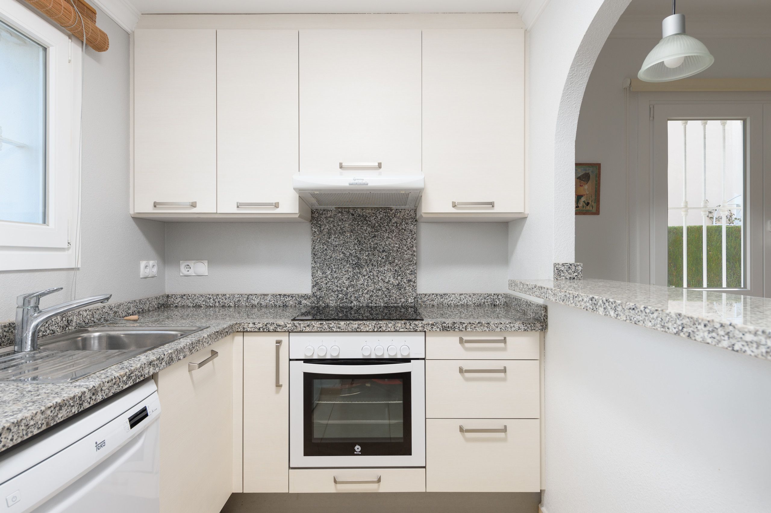 A modern kitchen with white cabinetry, granite countertops, a built-in oven, and a window with a view of a garden.