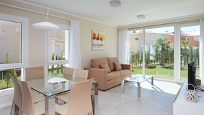 A modern living room with a glass dining table, beige sofa, and large windows offering a view of the garden and neighboring buildings. - Moss Rose by ON Travel (Oliva)