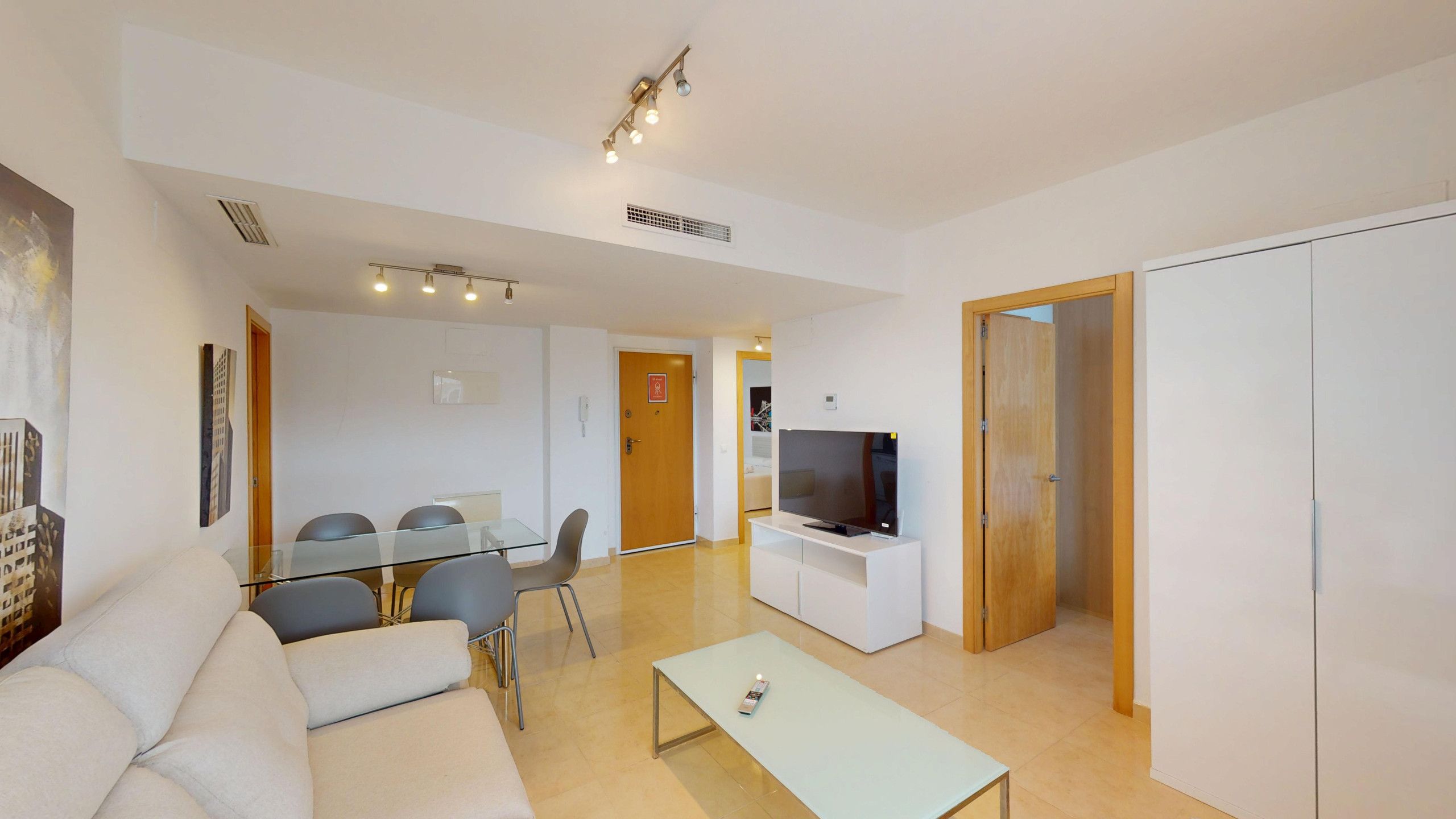 Una sala moderna con un sofá, una mesa de comedor de cristal con sillas, un televisor sobre un mueble bajo y un armario de madera.