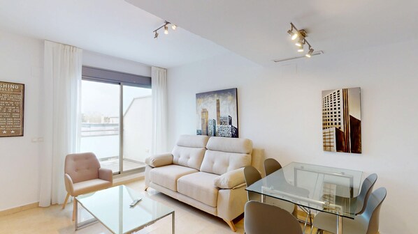 A modern living room with a sofa, a glass coffee table, and a dining table with chairs. There is a large window with curtains, a painting of buildings on the wall, and a ceiling light fixture. - Lily by ON Travel (Oliva)