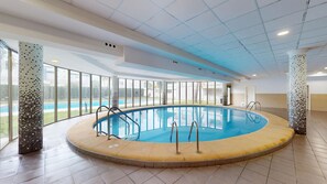 A large indoor swimming pool with clear glass walls, a tiled floor, and a view of a pool outside. - Lily by ON Travel (Oliva)