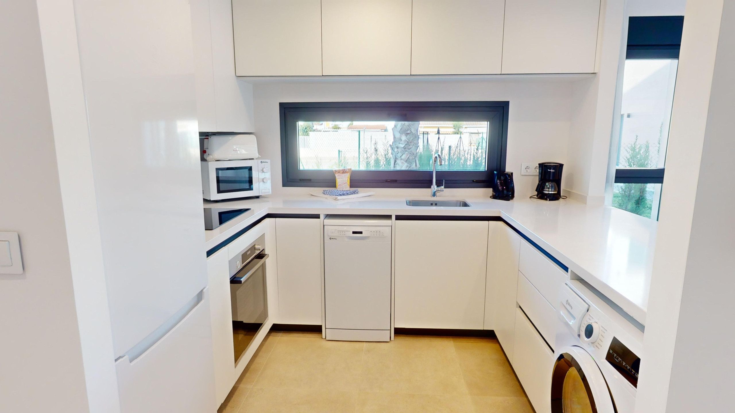 Een kleine keuken met witte kastjes, een vaatwasser, een magnetron, een koffiezetapparaat en een raam voor natuurlijk licht.
