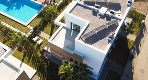Vista aérea de un edificio moderno con un panel solar en la azotea, una piscina y un césped bien cuidado con palmeras.