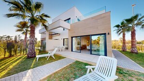 A modern two-story house with a patio area, white outdoor furniture, and palm trees.