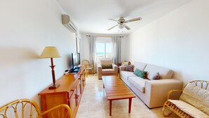 Ein helles Wohnzimmer mit einer Couch, einem Holztisch, einem Fernseher und einem Essbereich mit Stühlen und Tisch.