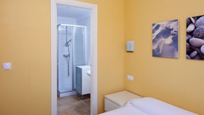 Un baño con ducha, inodoro y lavamanos. Incluye una cama con ropa blanca y una mesita de noche pequeña.
