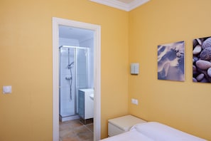 Ein Badezimmer mit Dusche, Toilette und Waschbecken. Ein Bett mit weißer Bettwäsche und ein kleiner Nachttisch sind ebenfalls vorhanden.