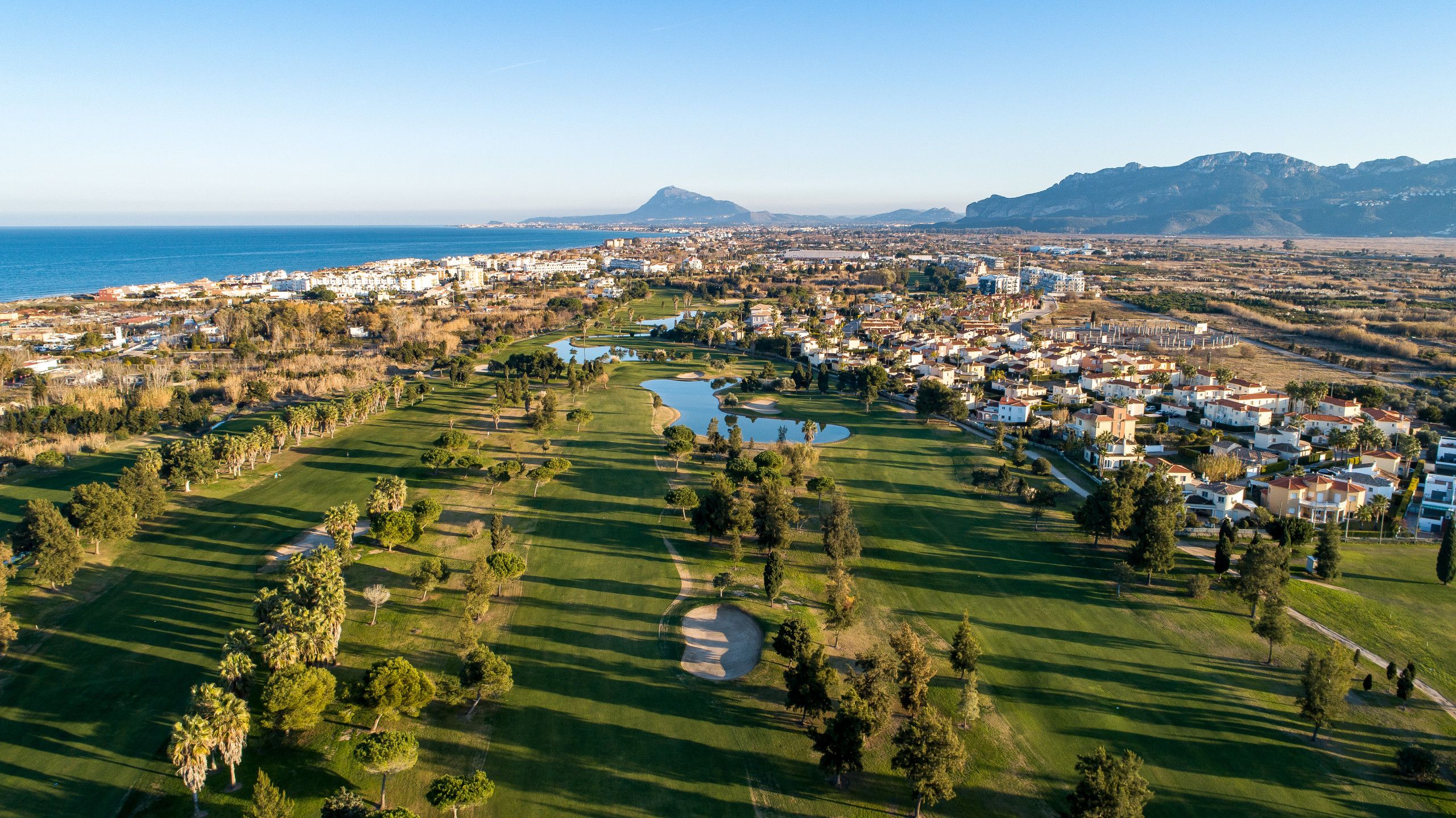 An aerial view of a coastal town with a golf course, residential areas, and a mountain in the background.
