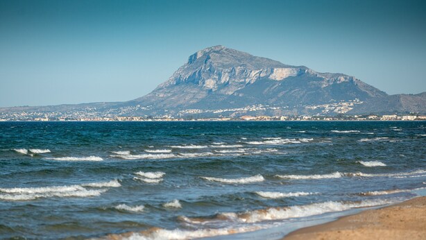 A beach with waves, a mountain in the background, and a clear sky. - Velvet by ON Travel (Oliva)