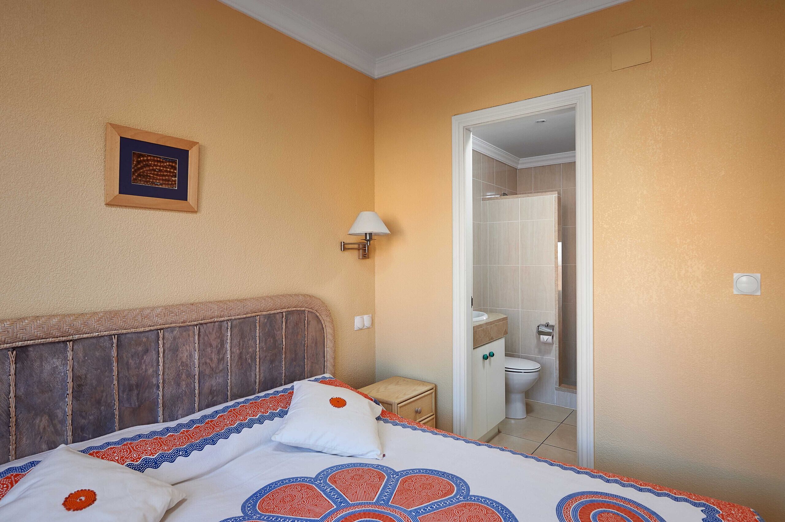 Ein Schlafzimmer mit Holzkopfteil, einem Bett mit gemusterten Betttextilien, einer Nachttischlampe und einem gerahmten Bild an der Wand.