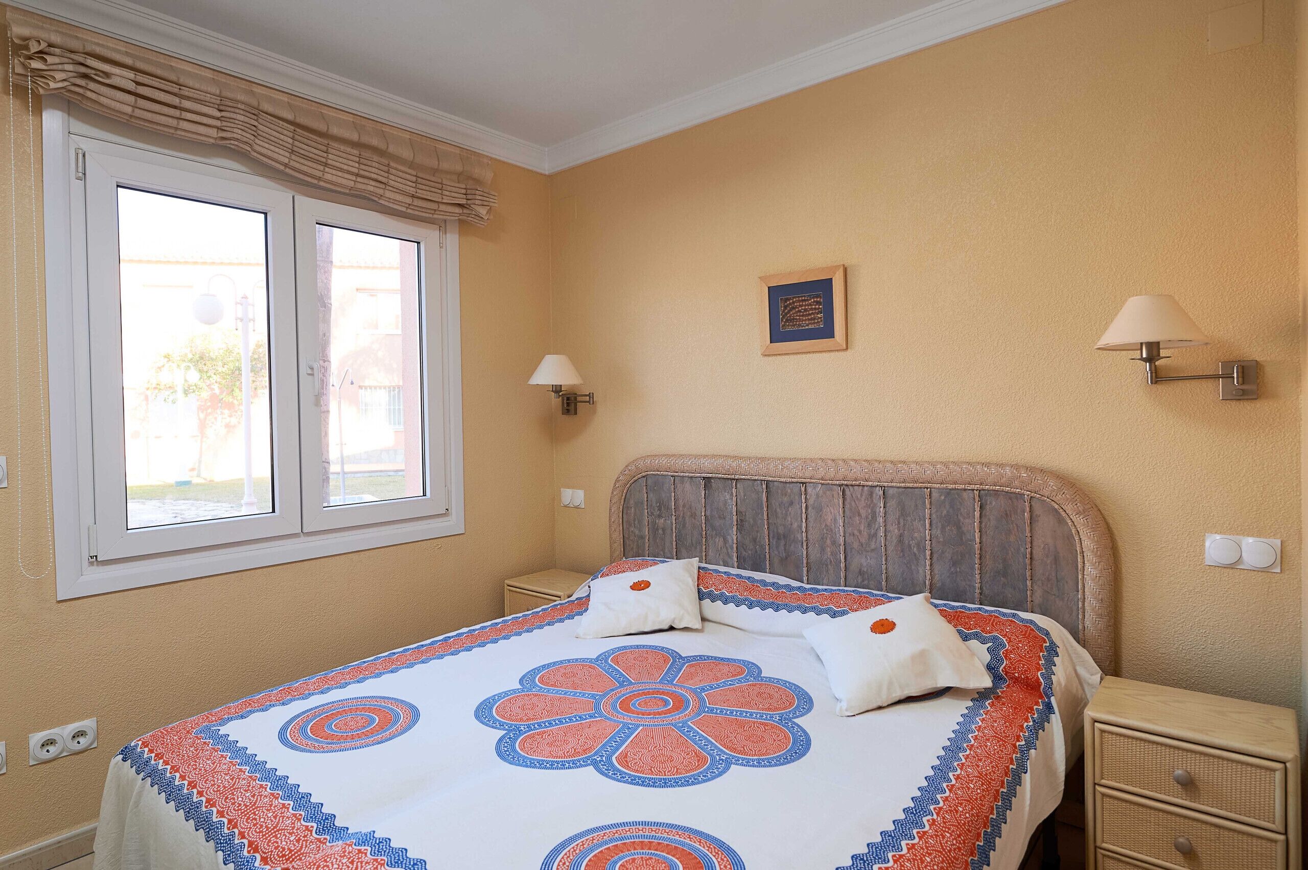 Une chambre à coucher comprenant un lit, deux lampes de chevet, une fenêtre avec des rideaux, une petite table de chevet et un tableau accroché au mur.