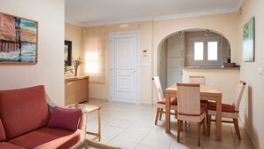 A living room with a red sofa, a dining table with wicker chairs, a kitchen area with a window, and a white door. - Yarrow by ON Travel (Oliva)