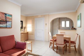 A living room with a red sofa, a dining table with wicker chairs, a kitchen area with a window, and a white door.