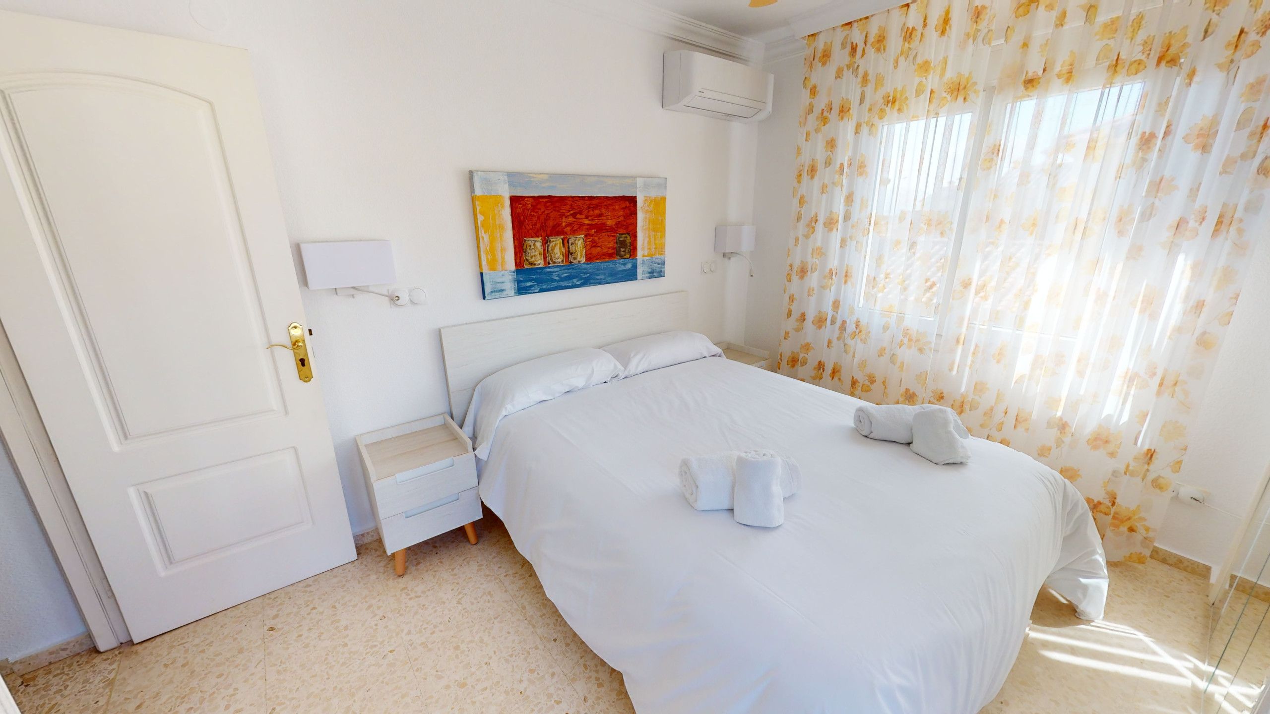Een hotelkamer met een bed, nachtkastje, wandairconditioning en een raam met bloemen gordijnen.