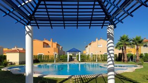 Een zwembad met een blauwe pergola erboven, omgeven door strakke hagen en palmbomen.