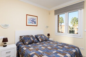 Un dormitorio con cama, dos mesitas de noche, una lámpara auxiliar, un cuadro enmarco y una ventana con vistas a palmeras.