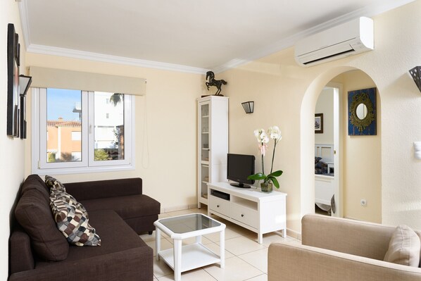 Una sala de estar con un sofá marrón, una mesa de centro blanca, un televisor sobre un mueble blanco y una orquídea en maceta.