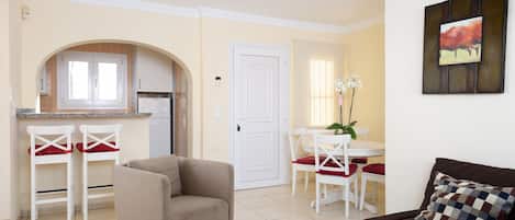 Una sala de estar con un sofá marrón y una mesa de centro de vidrio, un comedor con una mesa y sillas blancas, y una cocina con un refrigerador y una ventana.
