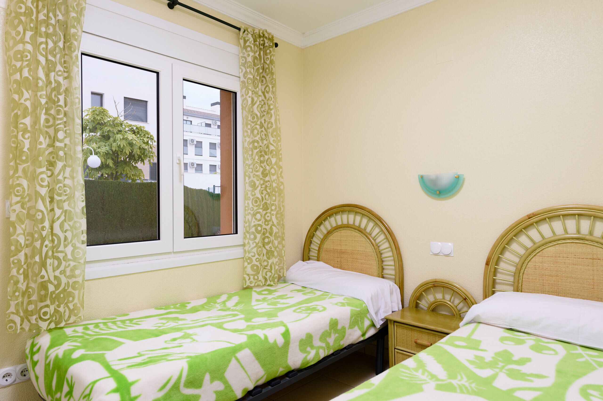 Une chambre avec deux lits simples, une fenêtre avec des rideaux, un luminaire fixé au mur et une vue sur les bâtiments à l’extérieur.