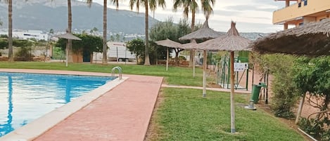 A swimming pool with a tiled walkway, surrounded by palm trees and a building with balconies.
