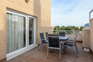 A balcony with a table and chairs, a sliding glass door, and a view of palm trees and buildings.