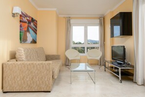 A living room with a sofa, two chairs, a coffee table, a flat-screen TV on a stand, a painting on the wall, and a window with curtains. - Denia Beachfront apartment by ON Travel (Denia)