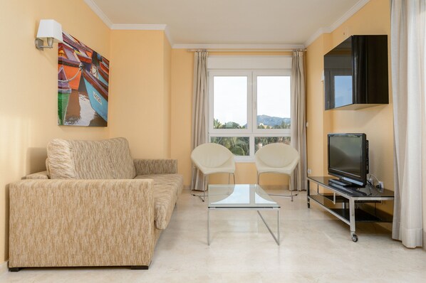 A living room with a sofa, two chairs, a coffee table, a flat-screen TV on a stand, a painting on the wall, and a window with curtains.
