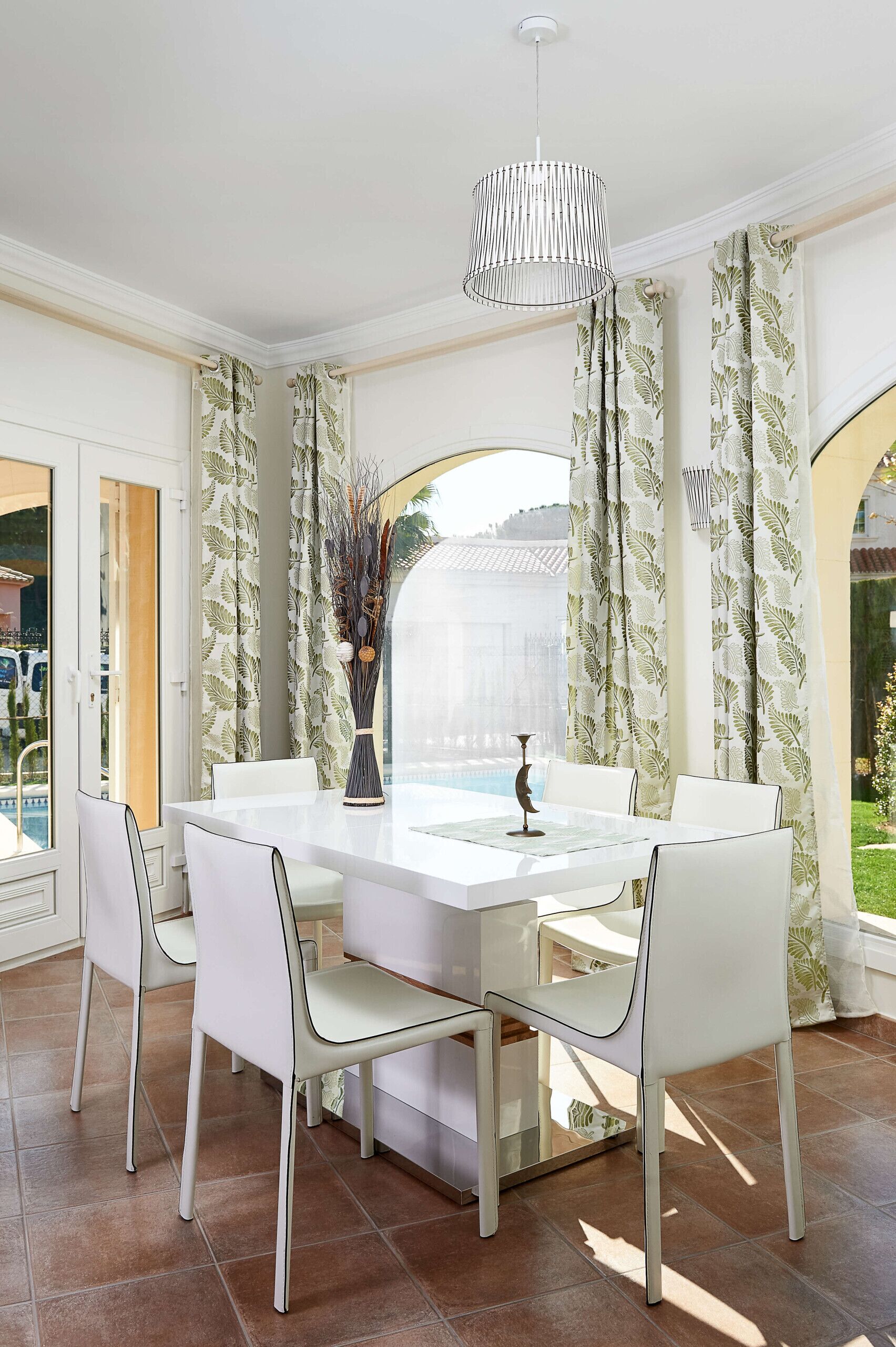 Een eethoek met een witte tafel en stoelen, een vaas met gedroogde planten en een hanglamp.