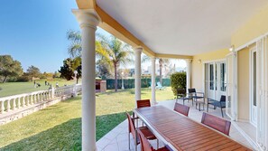 Un portico coperto con tavolo e sedie in legno, vista su un campo da golf e ringhiera bianca.
