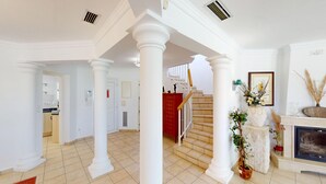 Un vestíbulo amplio con columnas blancas, una escalera, una chimenea y una zona de cocina visible a través de una puerta abierta.