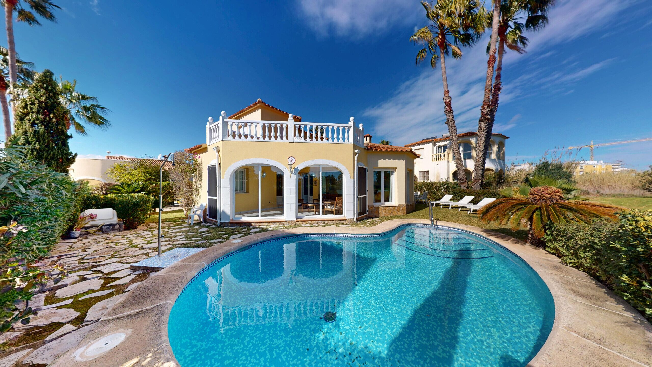 Una casa de dos pisos con piscina, rodeada de palmeras y un cielo azul despejado.