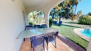 Eine Terrass mit Glastisch und blauen Stühlen, umgeben von einem Pool und viel Grün.