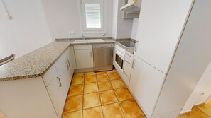 Una cocina compacta con armarios blancos, lavavajillas, horno y ventana con persianas.