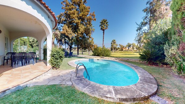A swimming pool with a stone deck, surrounded by grass and trees, and a patio area with chairs.
