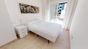 A bedroom with a white bed, a nightstand with drawers, a painting on the wall, and a view of a building through a glass door.